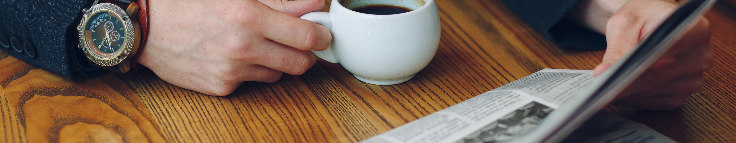 Man's hands close-up holding cup of coffee and a newspaper on a wood table. Businessman in costume and vintage watch reading news while holding a white cup of coffee.