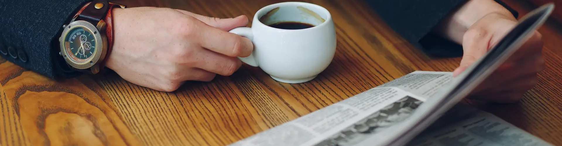 Man's hands close-up holding cup of coffee and a newspaper on a wood table. Businessman in costume and vintage watch reading news while holding a white cup of coffee.